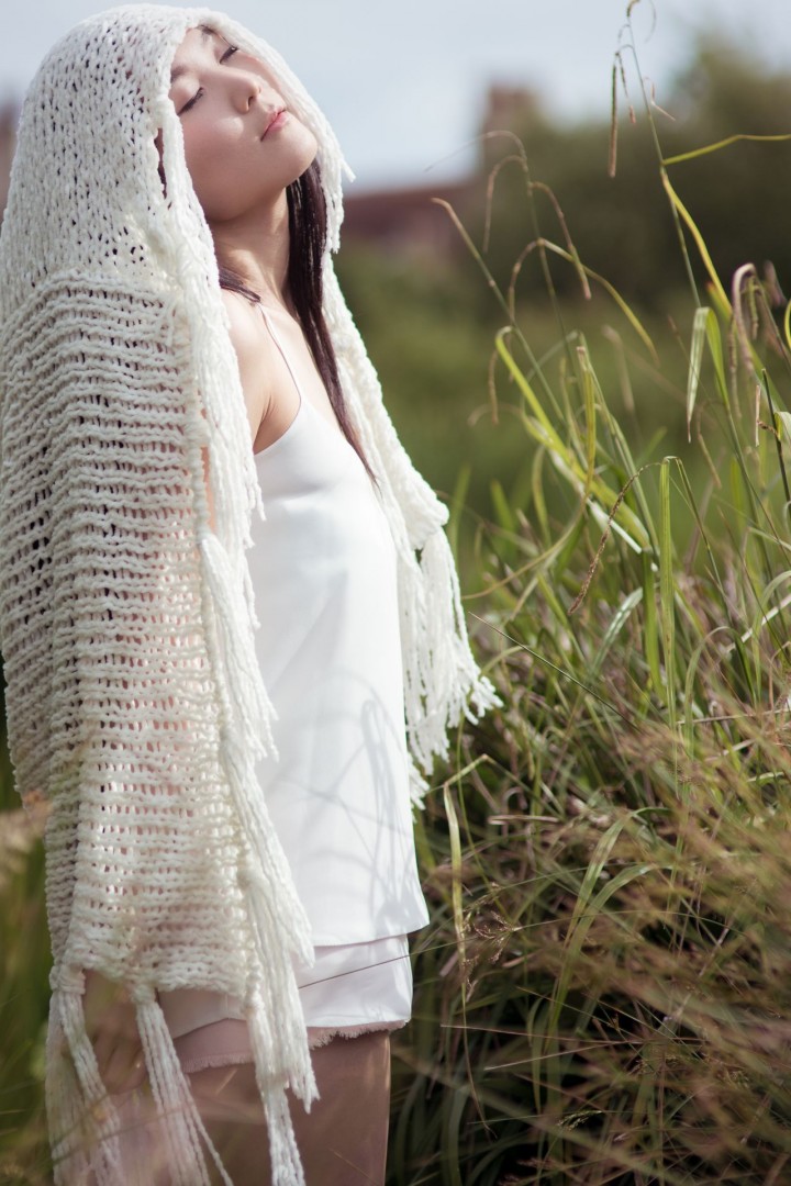 model in white top and shawl with eyes closed in bright sun beside tall grasses